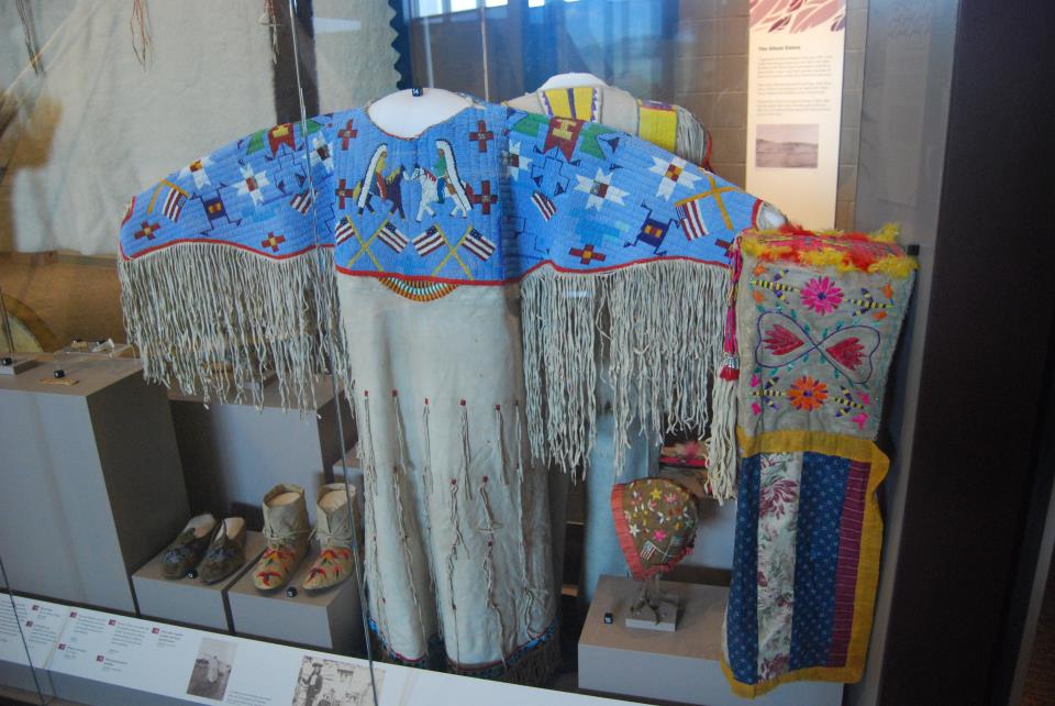 A native dress with blue quillwork on the chest and upper back portion that expands into the arms. Depicted in the blue section are native americans in headdresses riding horses, American flags, and other symbols