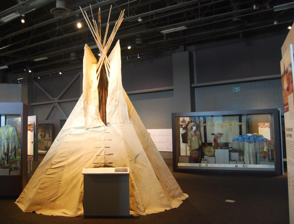 A tan colored tipi stands on exhibit in a gallery