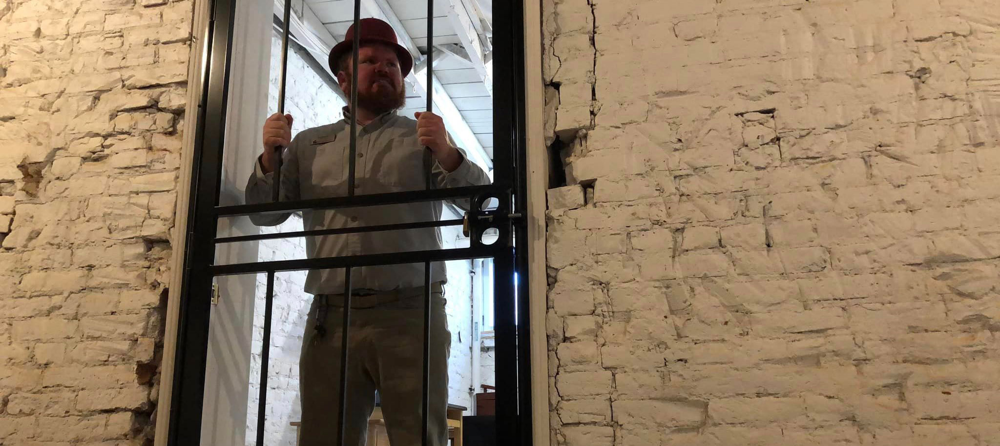 A man in a hat, button up shirt, and pants with belt stands behind a jail door.