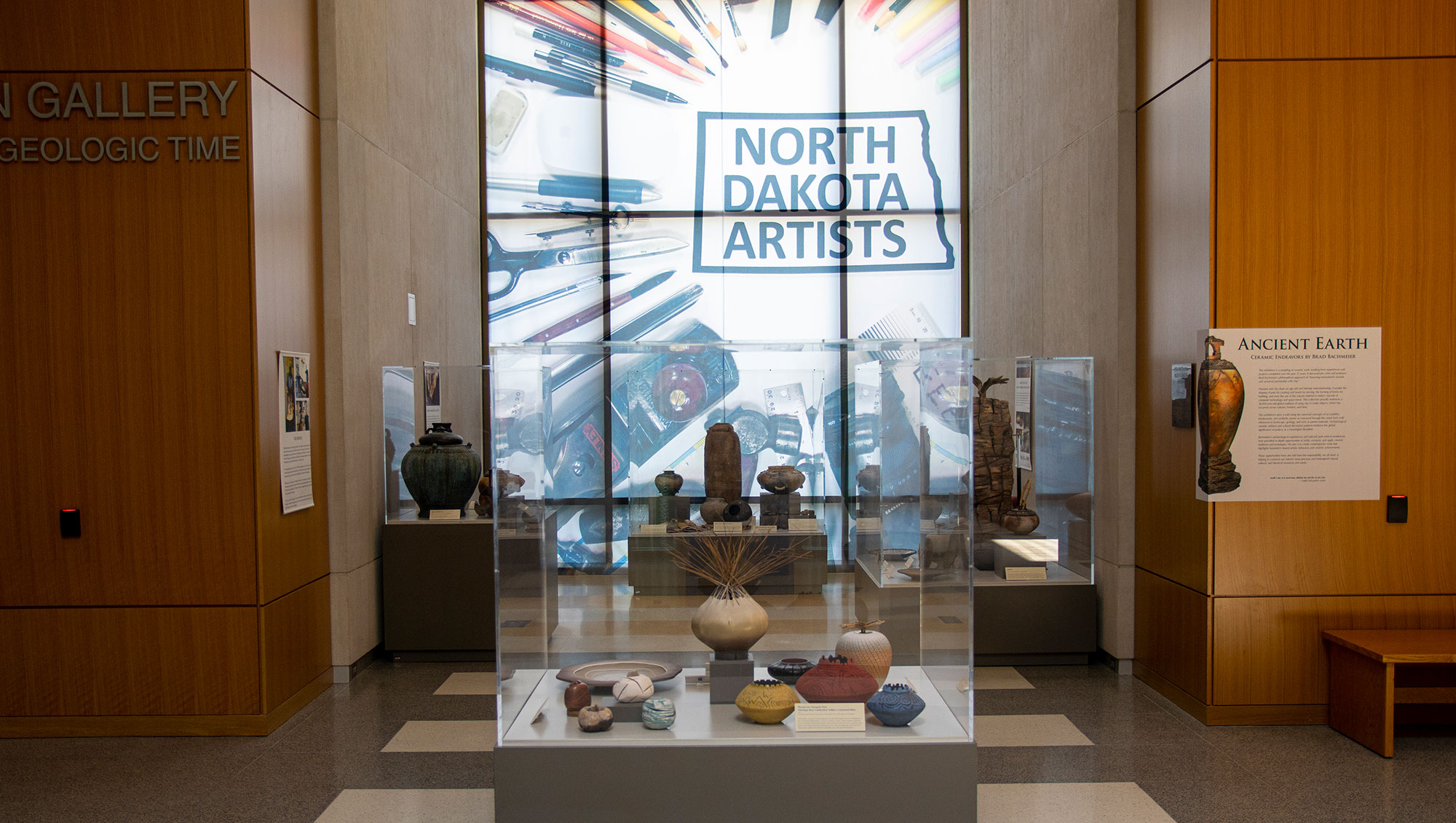 Hallway with display cases containing pottery. In the background is a window with a window cling that reads North Dakota Artists and has art supplies around the text
