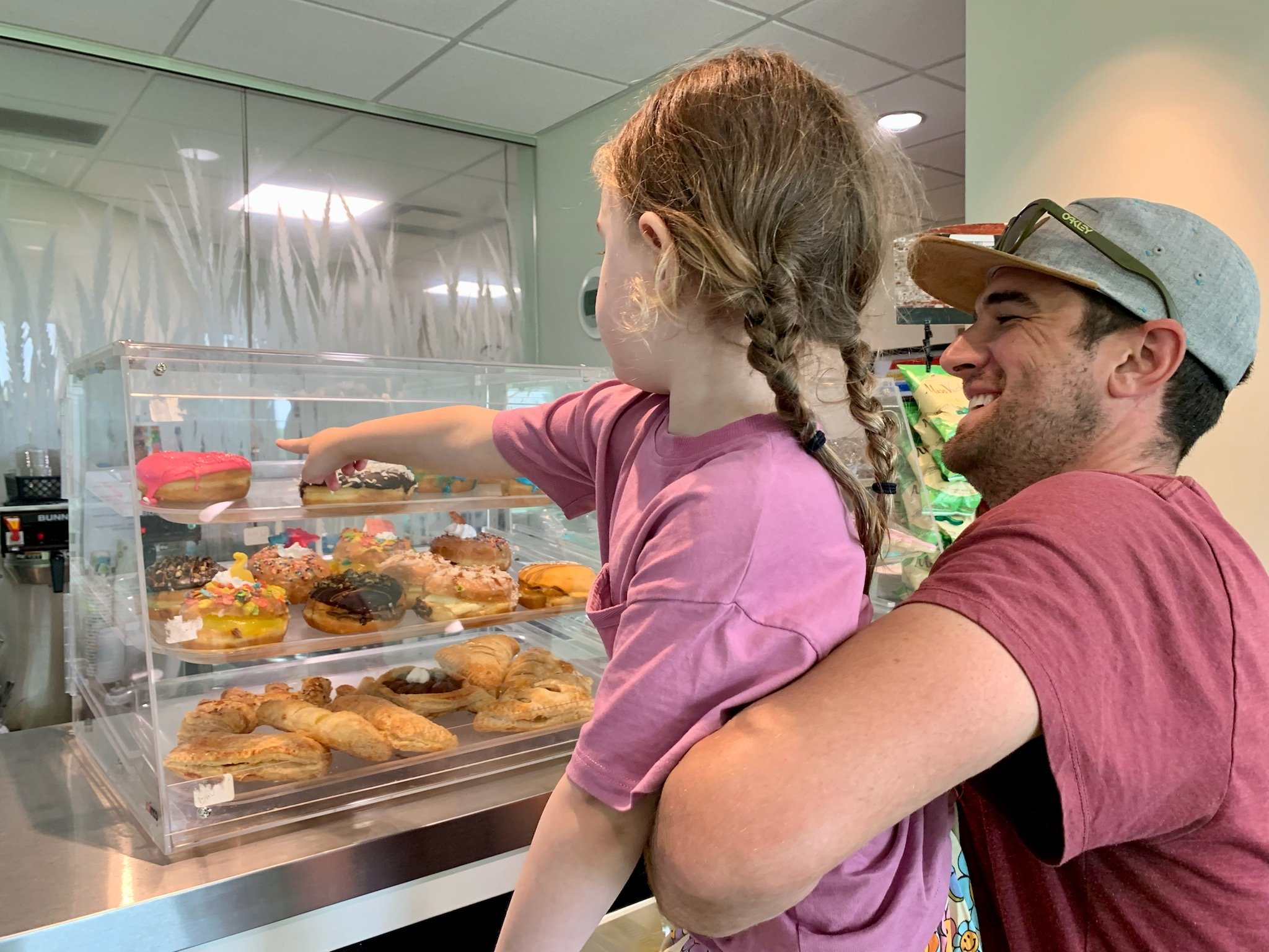 A dark haired man with stubble wearing a red t-shirt and gray hat with Oakley sunglasses sitting on the hat holds up his young daughter with braided low pigtails and a pink shirt to show her what's in the dessert display. The girl points to a donut with pink frosting.
