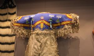 A Native American dress with blue, yellow, and red beading at the top and tassles hanging down from the arms
