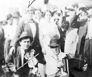 A man wearing a hat and suit jacket plays a violin while the man next to him in a hat, button up shirt, and tie plays an accordion. A group of people stand behind them with some of the men drinking bottles of beer.