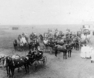 Many horses and buggies are shown with roughly 4 passengers per buggy. The front one carries a bride and groom. On the right side of the image are 7 people standing dressed in white.