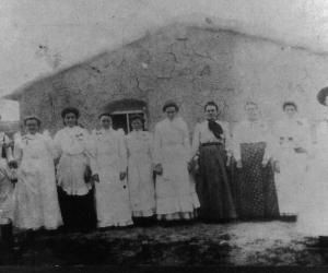 8 women and 2 men who are on either end stand in front of a summer kitchen. White aprons and ribbons identify the attendants for the reception. The two stewards hold schnapps bottles and wear women’s bib aprons. Several of the women sport red bows.