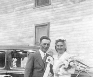 A bride and groom stand in front of a car. The groom has ribbons hanging on his lapel.